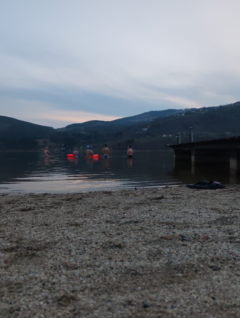 Vier Menschen stehen in der Dämmerung mit orangen Schwimmbojen im kalten Wasser des Stubenbergsees